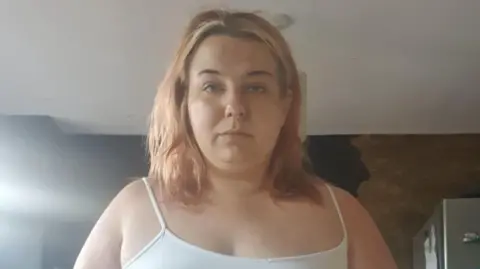 Sammi Hill Sammi Hill, a woman in her thirties. She is pictured wearing a white vest and is looking at the camera. She weighed 18 stone in this image.