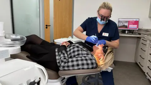 Dr May Bassett treating a patient lying in a dentist's chair. May is wearing a navy scrubs, with a white name badge. Her blonde hair is tied back and she wears a pale blue mask and special glasses with attachments that allow a magnified view of the patient's teeth. Her gloved hands are holding an instrument in the open mouth of a patient. The patient is wearing dark glasses and has short blonde hair. She is wearing a black and white checked jacket over a black dress, with black tights. Surrounding them are the cupboards and equipment of a dentist's treatment room.