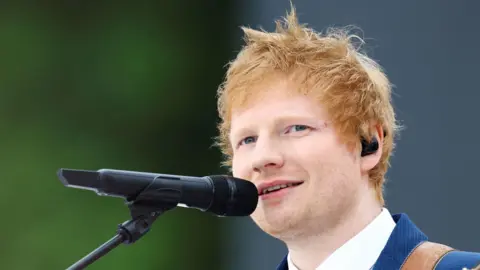 PA A close-up photo of Ed Sheeran talking into a microphone. He is wearing a white shirt with a blue blazer over the top of it. He is wearing a black earpiece. 