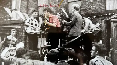 Charlie Roberts A black and white photo of The Quarrymen playing in front of a house. There is a drummer and multiple guitar players in the six-strong group. A young John lennon sings into a microphone. The image has been changed to colourise him. He has short brown hair and wears a red check shirt.