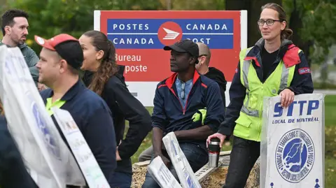 Bloomberg via Getty Images Striking postal workers, some holding white and blue picket signs reading 'STTP, local de Montreal', gather in front of a large blue and red Canada Post post office sign.