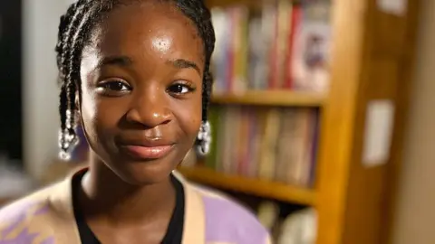 Martin Giles/BBC Hephzibah Akinwale who has dark hair in short braids pulled back from her face and is starting to smile. She is wearing a pink and yellow cardigan over a black top. Behind her is a bookshelf full of books.