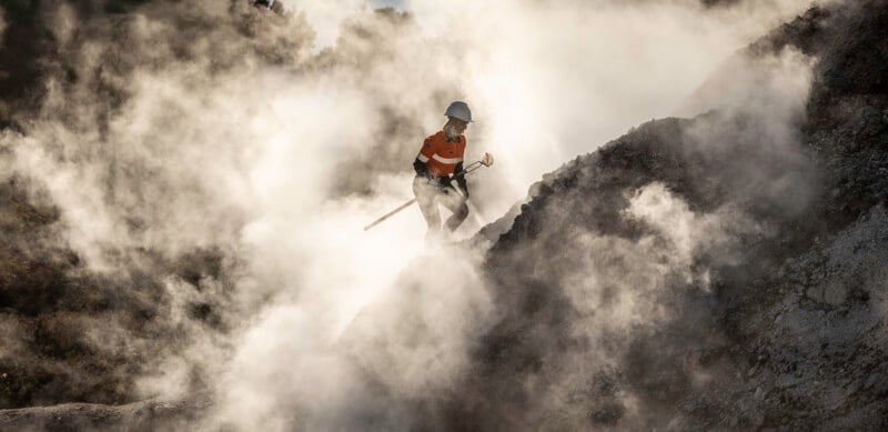 A person wearing a helmet and orange shirt stands on rocky terrain surrounded by thick clouds of white smoke or steam, holding a tool and appearing to work in a hazardous environment.