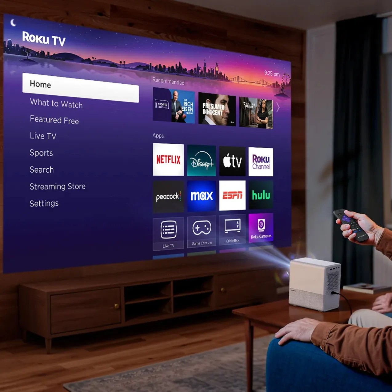 A Roku TV interface is projected on a wall, showing a home screen with menu options on the left and app icons including Netflix, Disney+, Apple TV, Roku Channel, Peacock, Max, ESPN, and Hulu in the center. A small white and gray projector is placed on a wooden coffee table, projecting the image. A person sitting on a blue couch is holding a Roku remote, controlling the projector. The room has wooden flooring, a wooden media console under the projection, and dark curtains on the right side.