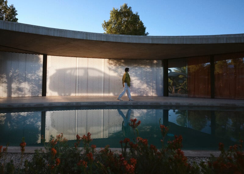 A person in a yellow shirt and white pants walks by a modern building with curved architecture, reflected in a calm pool, with greenery and orange flowers in the foreground and trees in the background.