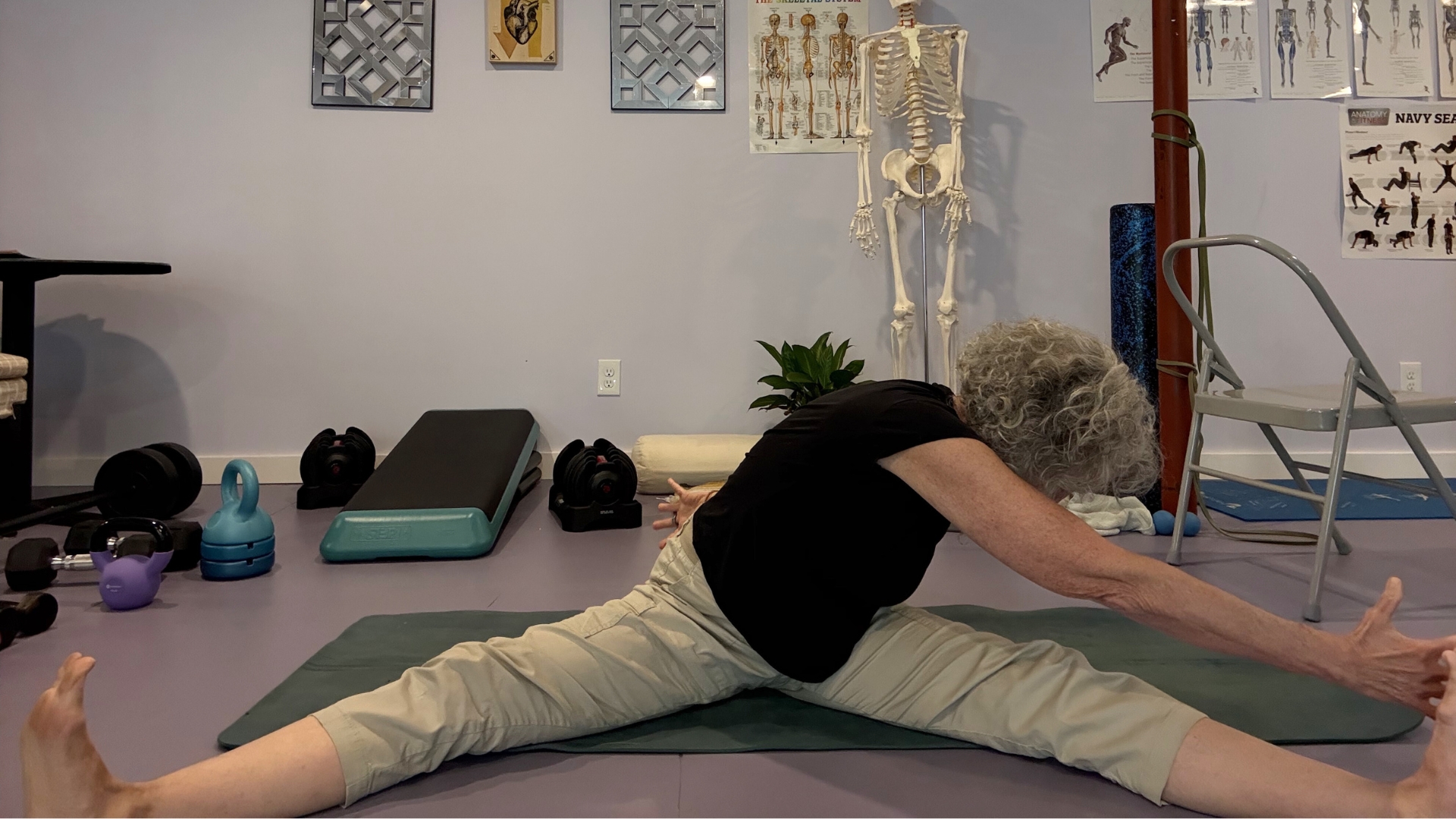 woman in light trousers and dark top sat on the floor legs outstretched wide on a green mat in a studio setting, with one hand reaching to her opposite foot.