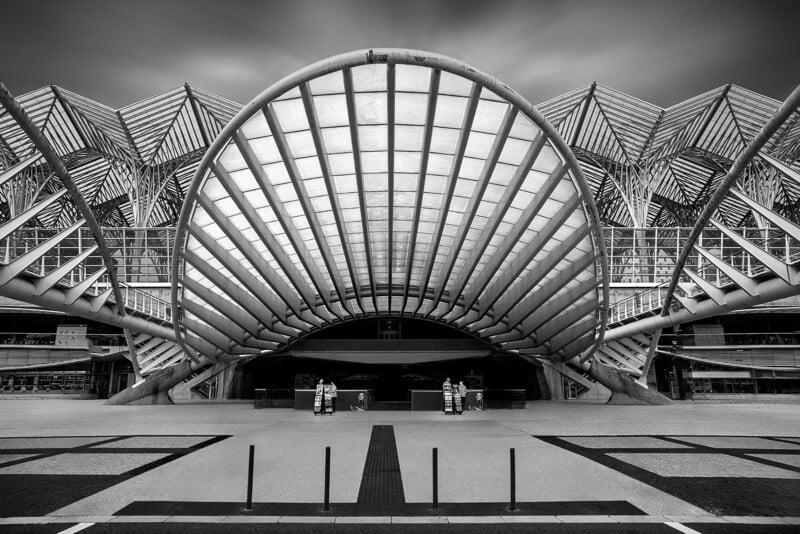 A striking, modern architectural structure with a large, curved, ribbed canopy and geometric patterns overhead, captured in black and white. Two people stand at the entrance beneath the sweeping design.
