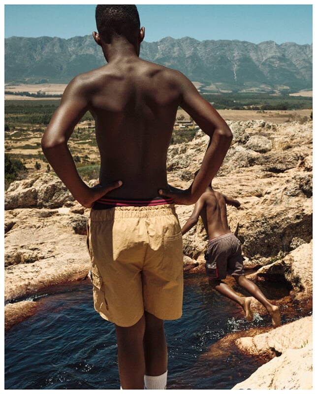 Two boys with dark skin are at a rocky pool; one stands with hands on hips, wearing tan shorts and white socks, while the other is mid-jump into the water. Mountains and fields stretch in the background.