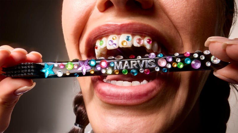 A person smiles with colorful rhinestones attached to their teeth, holding a black toothbrush decorated with similar rhinestones and the brand name "MARVIS.