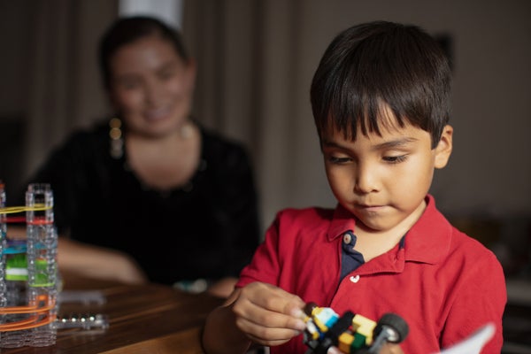 Young boy inred shirt plays with toy as mother in background looks on smiling
