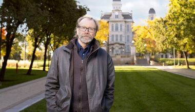 George Saunders, man in grey jacket standing on campus with Hall of Languages and fall foliage in the background