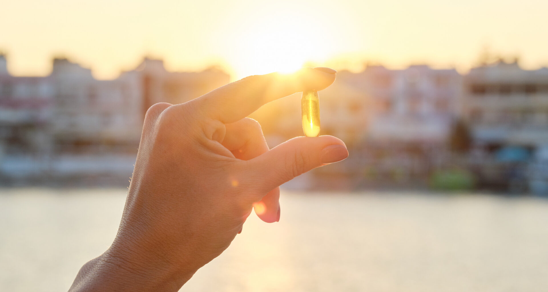 Hand holding translucent yellow pill against sun setting in background