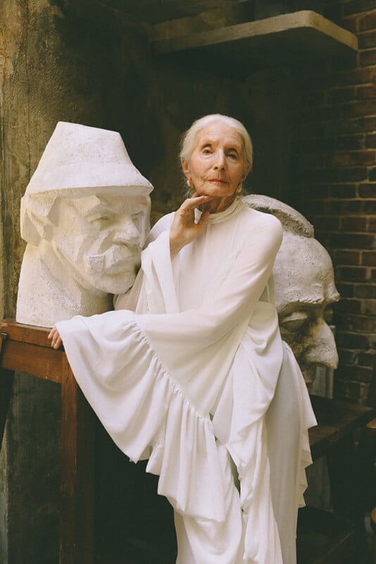 An elderly woman in a flowing white dress stands thoughtfully beside two large sculpted heads made of stone, set against a rustic background of brick and stone.