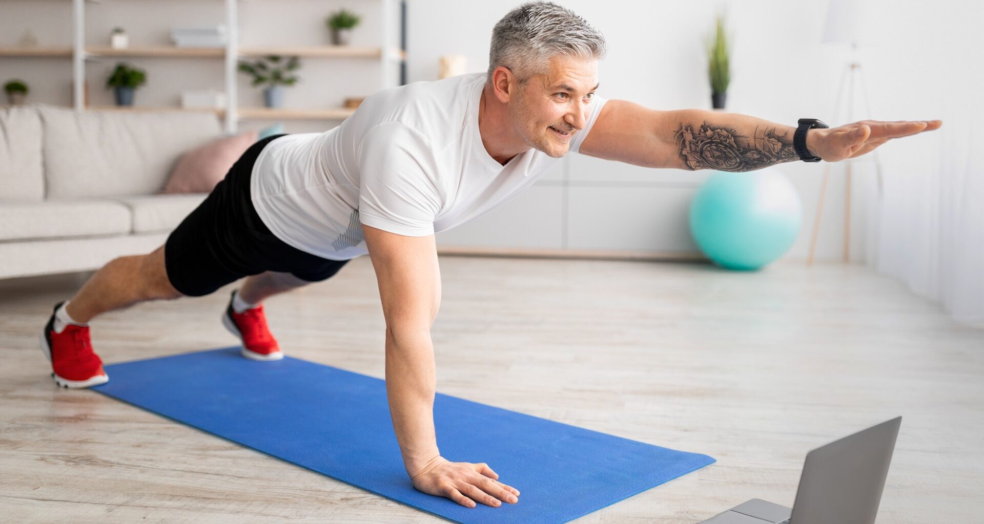 Older man performing one-arm plank on exercise mat at home during ab workout