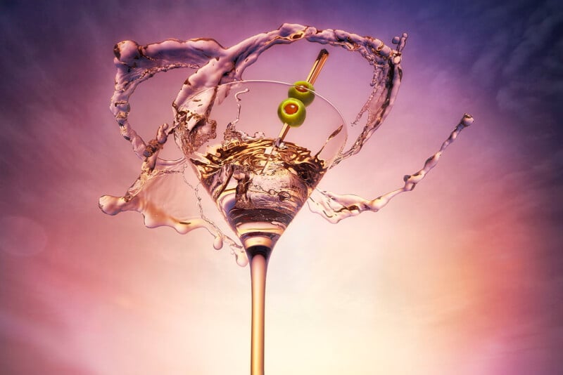 A martini glass with two green olives is splashed dramatically, with liquid swirling around it against a colorful, vibrant sky background.