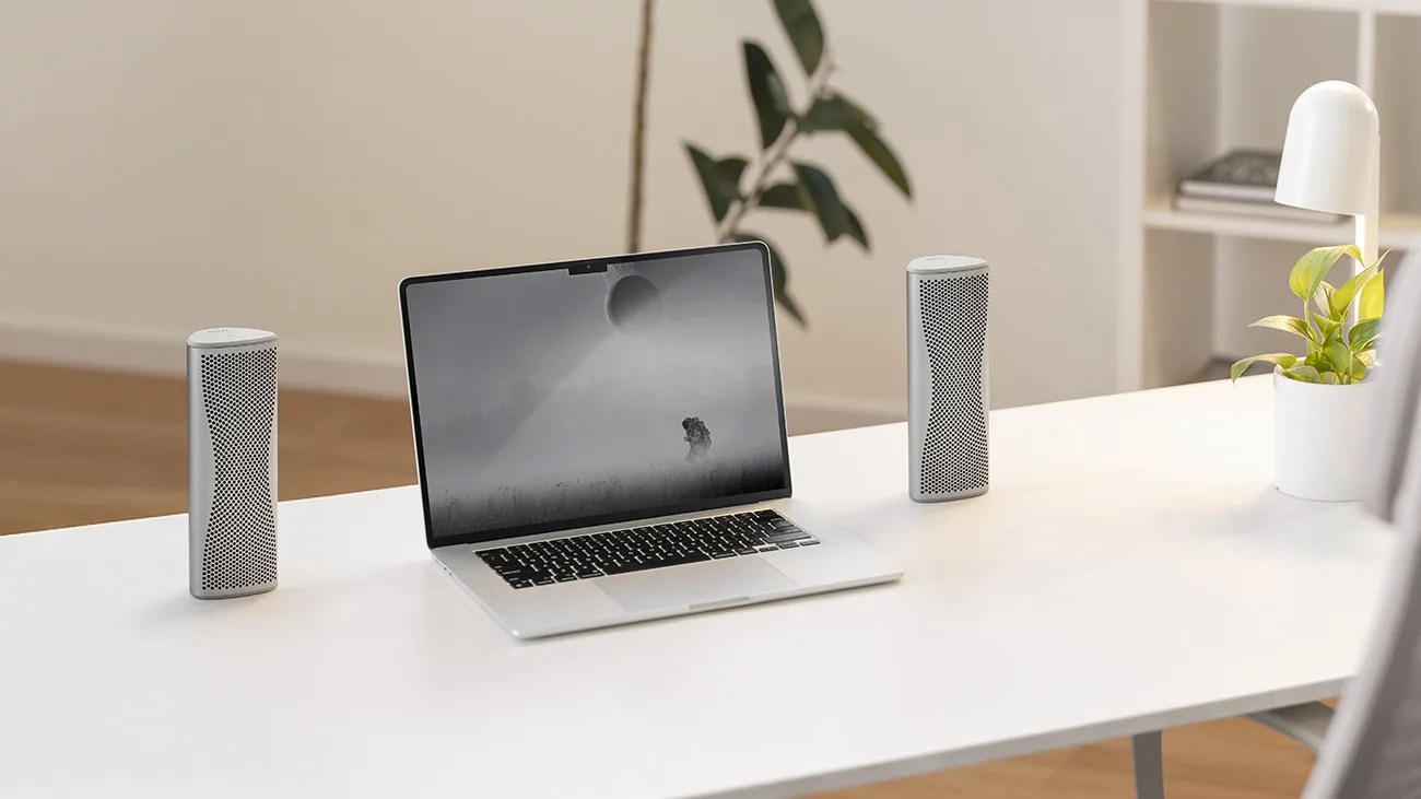 A silver laptop with a black and white screen displaying a misty landscape and a person standing in a field, placed on a white desk. On either side of the laptop are two identical silver speakers with a perforated front design. To the right of the desk is a white lamp and a white pot with a green plant. In the background, there is a blurred plant and a white shelving unit with books.