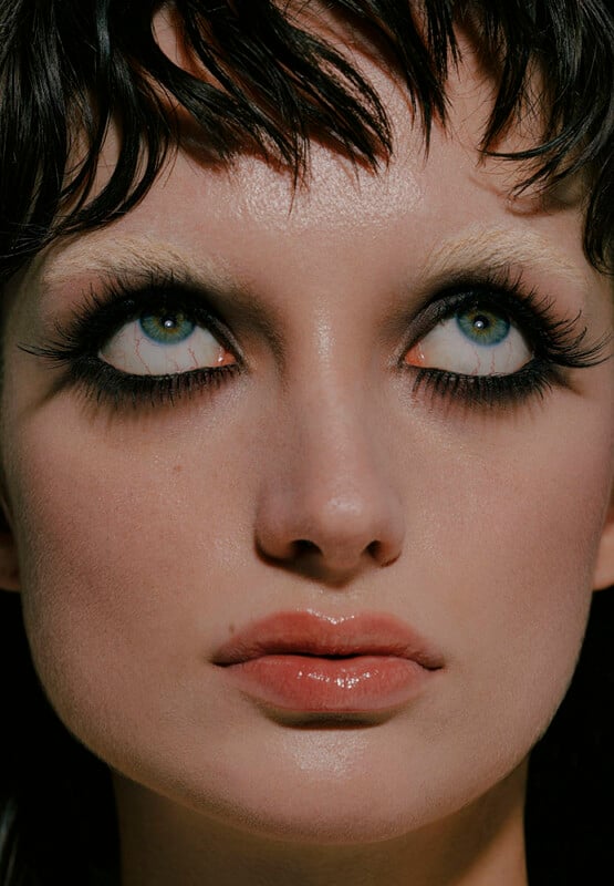A close-up of a person with light eyes looking upwards. They have dramatic black eye makeup, long lashes, light-colored eyebrows, glossy lips, and short, dark, wet-look hair with bangs.