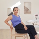 A woman sits on a wooden chair looking at the camera.