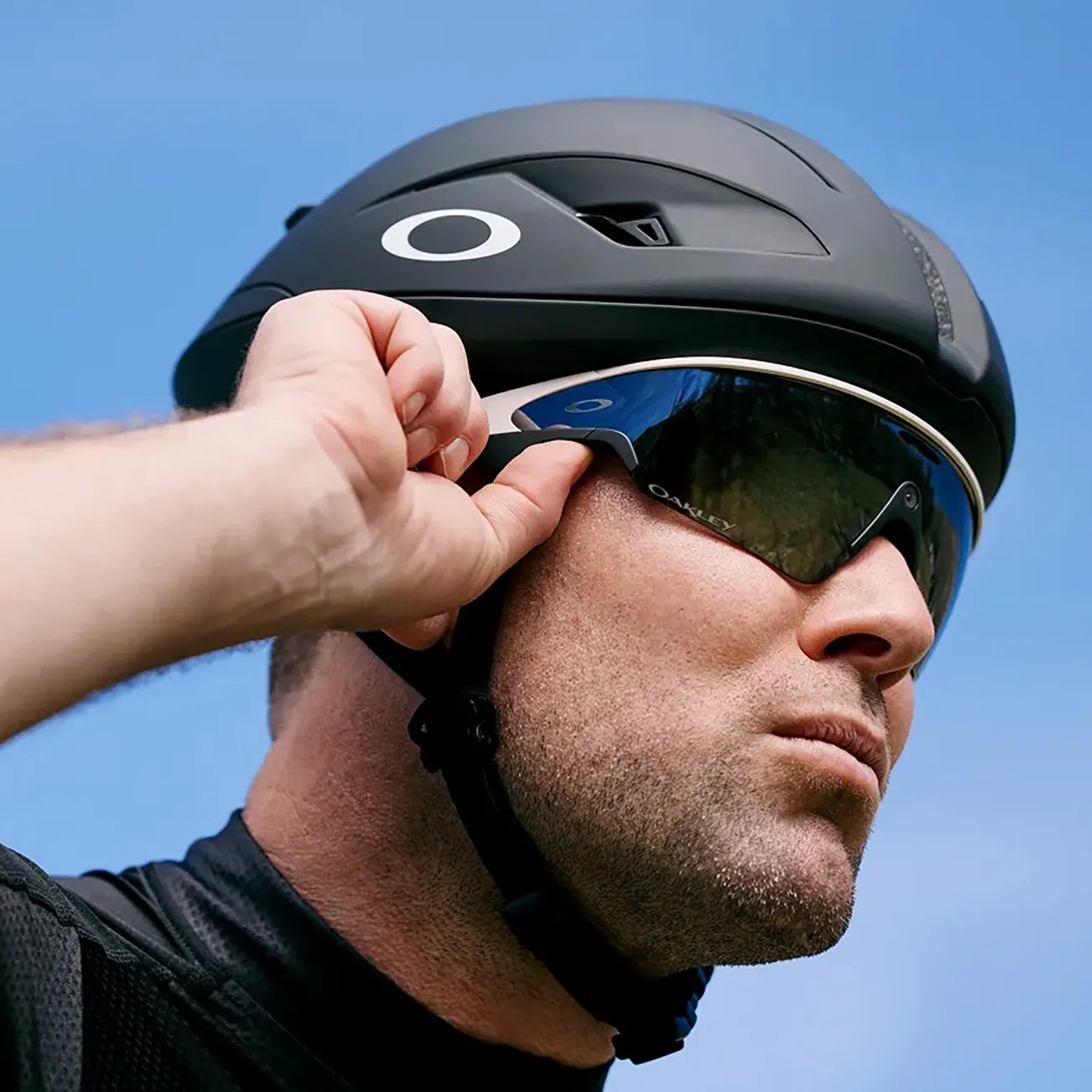 Close-up of a man wearing a black cycling helmet and reflective Oakley sunglasses, adjusting the sunglasses with his right hand against a clear blue sky background.