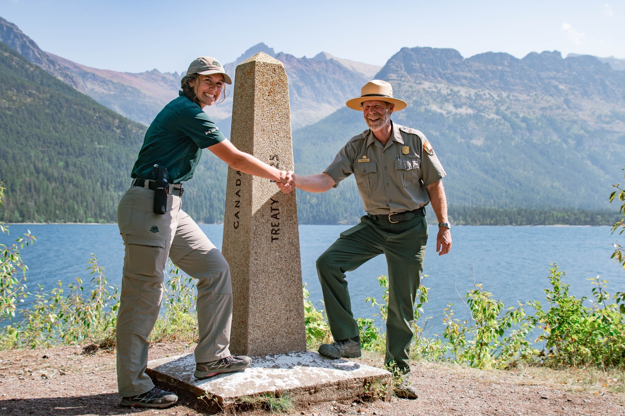 A Canadian and an American park warden shake hands across the border at the Waterton-Glacier International Peace Park. In the background, a blue lake and mountains.