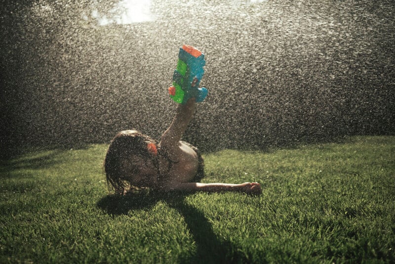 A child lies on the grass, holding up a colorful water gun, surrounded by a spray of water in the sunlight, enjoying outdoor summer play.