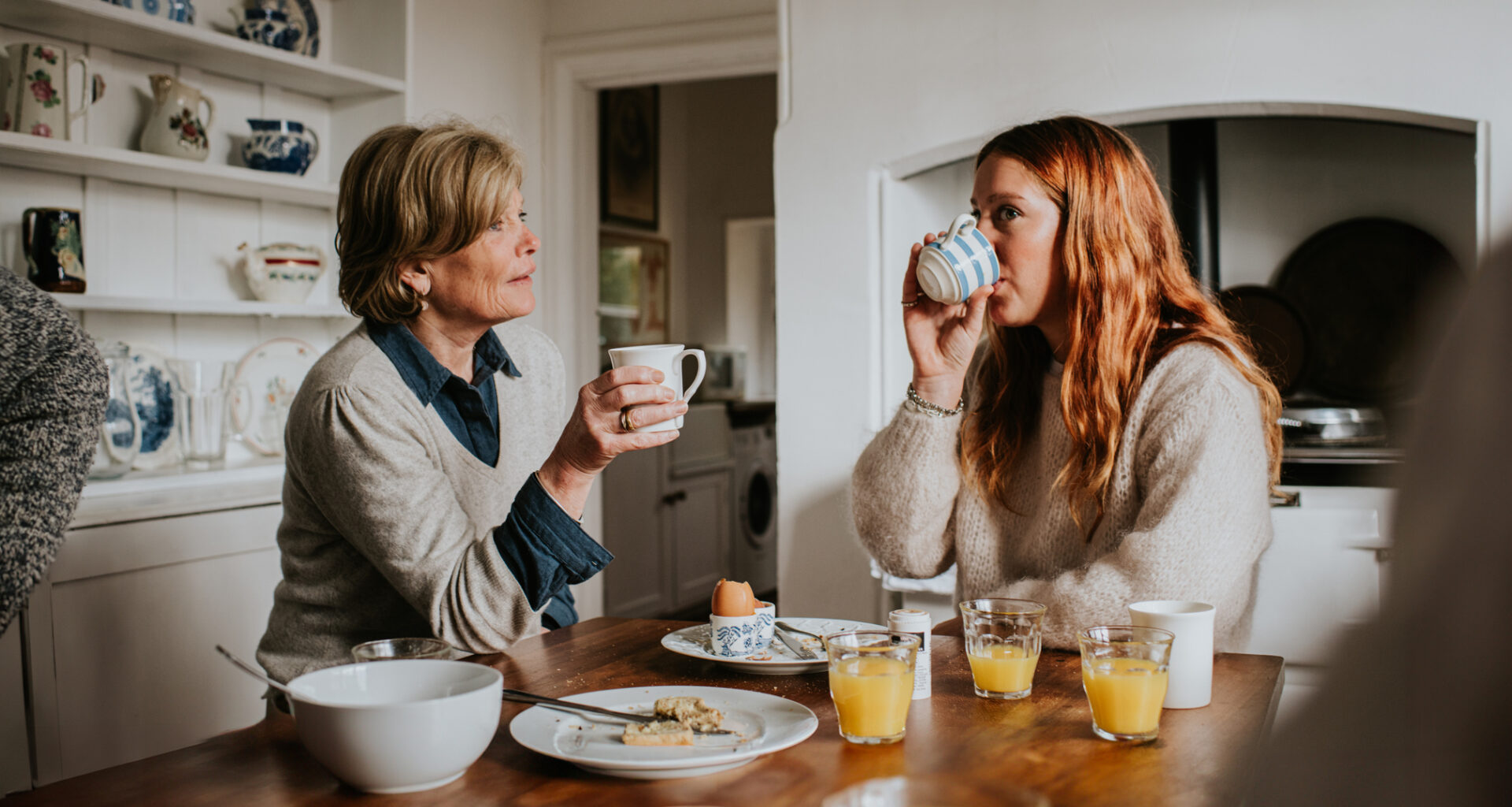 Two women sit at a table drinking from mugs, with eaten boiled eggs and glasses of orange juice on the table