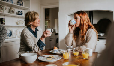 Two women sit at a table drinking from mugs, with eaten boiled eggs and glasses of orange juice on the table