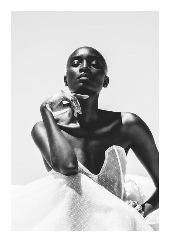 A black and white portrait of a woman with short hair, wearing a strapless, sheer polka-dot dress, sitting with one hand gracefully touching her chin, gazing confidently, with strong light and shadow contrast.