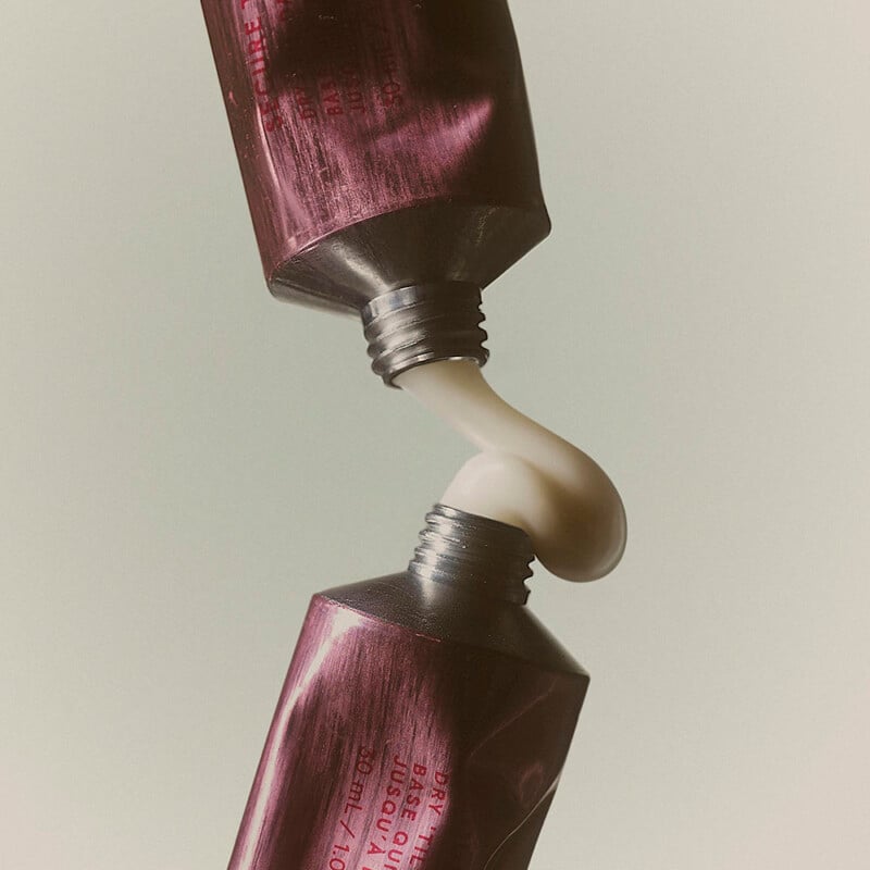 Two metallic tubes with their openings facing each other squeeze out white cream, which forms a smooth curve between them. The background is plain and neutral.