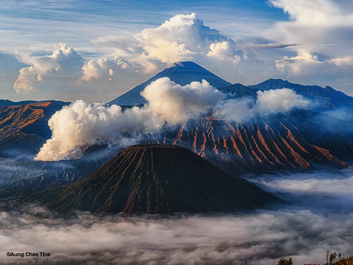 A scenic view of Mount Bromo and Mount Semeru in Indonesia at sunrise, with volcanic smoke rising, dramatic clouds, and mist covering the lower landscape.