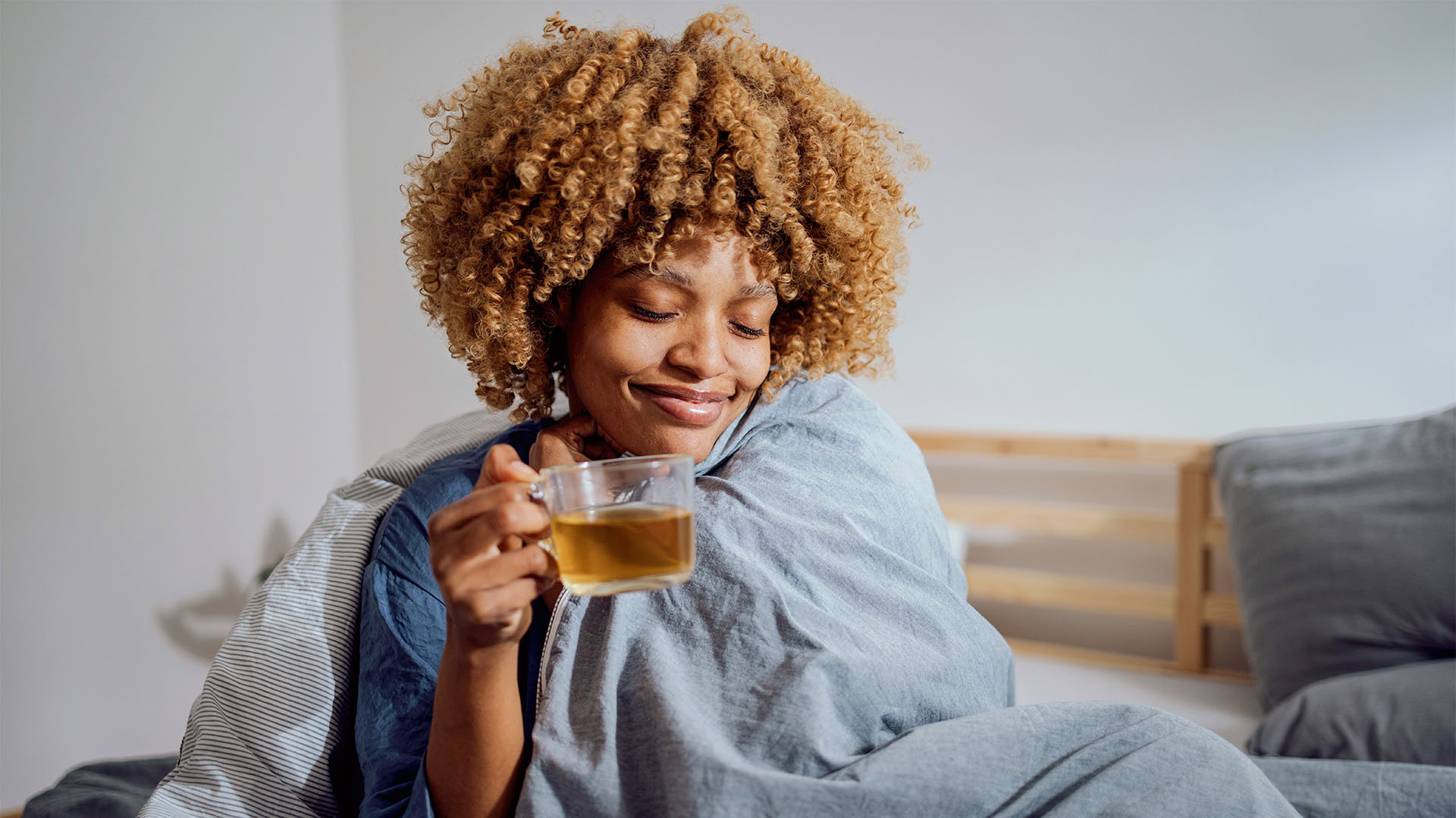 A woman sits on her bed wrapped in a blanket and holding a cup of green tea