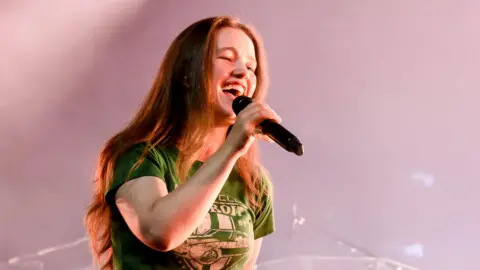 Getty Images Sigrid is singing into a mic smiling. She has long light brown hair, a green t-shirt on and denim jeans. Behind her on the stage is a blond woman playing the bass guitar. There is also a drum set behind her. 
