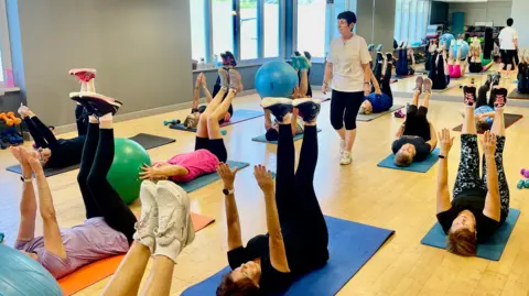 A woman with short black hair, black leggings and a white t-shirt is walking through a gym space. She is surrounded by twelve women laid on gym mats holding their legs and arms in the air. There is a full length mirror wall at the far end of the room and windows to the left. The woman laid on the floor are all wearing trainers and gymwear.
