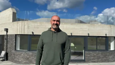 BBC A smiling man wearing a green hoody, standing in front of a black and white building. 