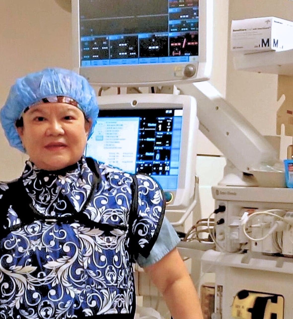 Woman wearing medical hair net and scrubs in front of monitors. 