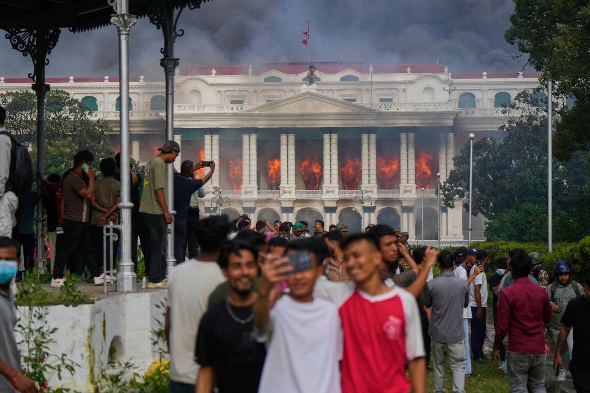 Three young males are shown gathered around a phone in the foreground as a long, august-looking building is on fire in the background.