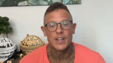 Robert speaking to camera in an interview. He wears glasses and a pinkish top and is sitting in front of some of his trophies.