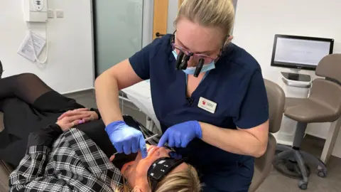 BBC May is wearing her navy blue, short-sleeved scrubs and special glasses with a light and magnifier capacity to inspect a patient's teeth. She is wearing a face mask and blue gloves and is inspecting the teeth of a patient lying on the chair in front of her. They are in a modern dental practice with a chair and computer visible in the background.