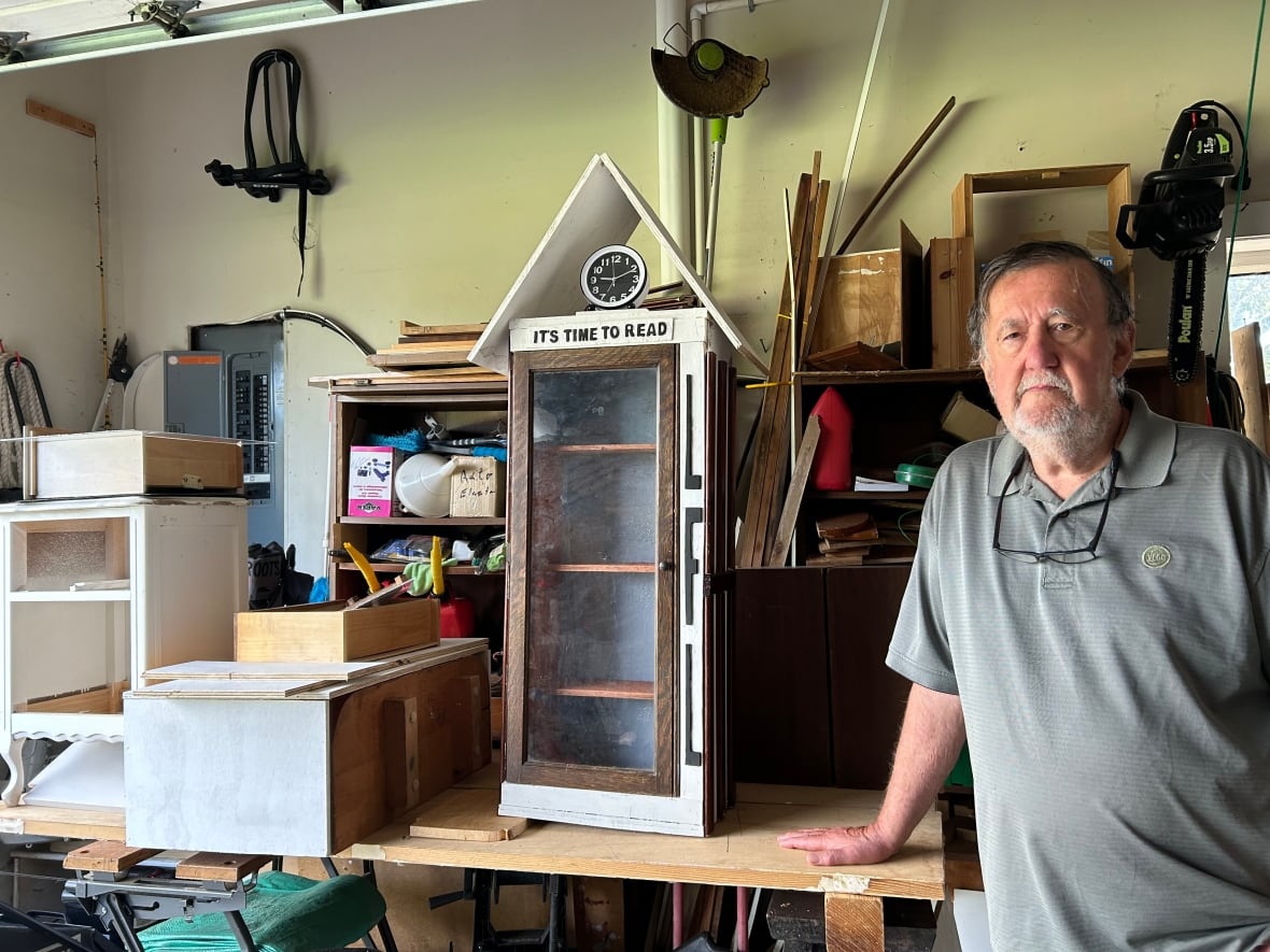 A senior man in a grey T-shirt stands in a garage with two little free libraries.