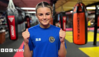 A girl with blonde-streaked hair, wearing a blue t-shirt, smiles at the camera. Behind her are punchbags