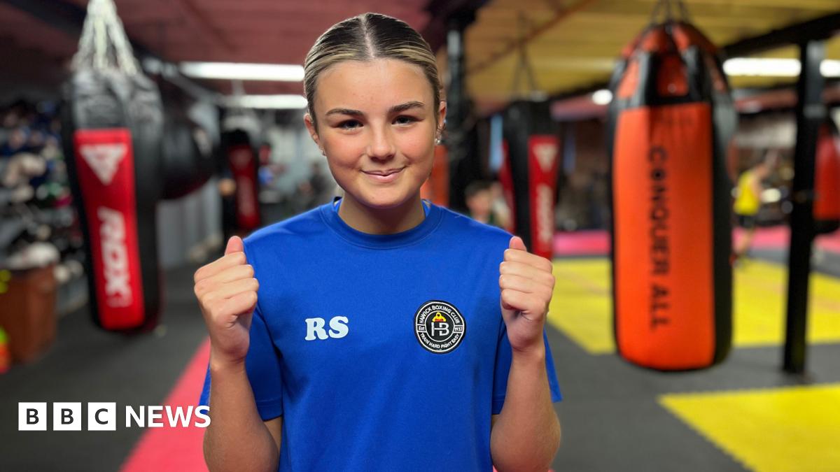 A girl with blonde-streaked hair, wearing a blue t-shirt, smiles at the camera. Behind her are punchbags