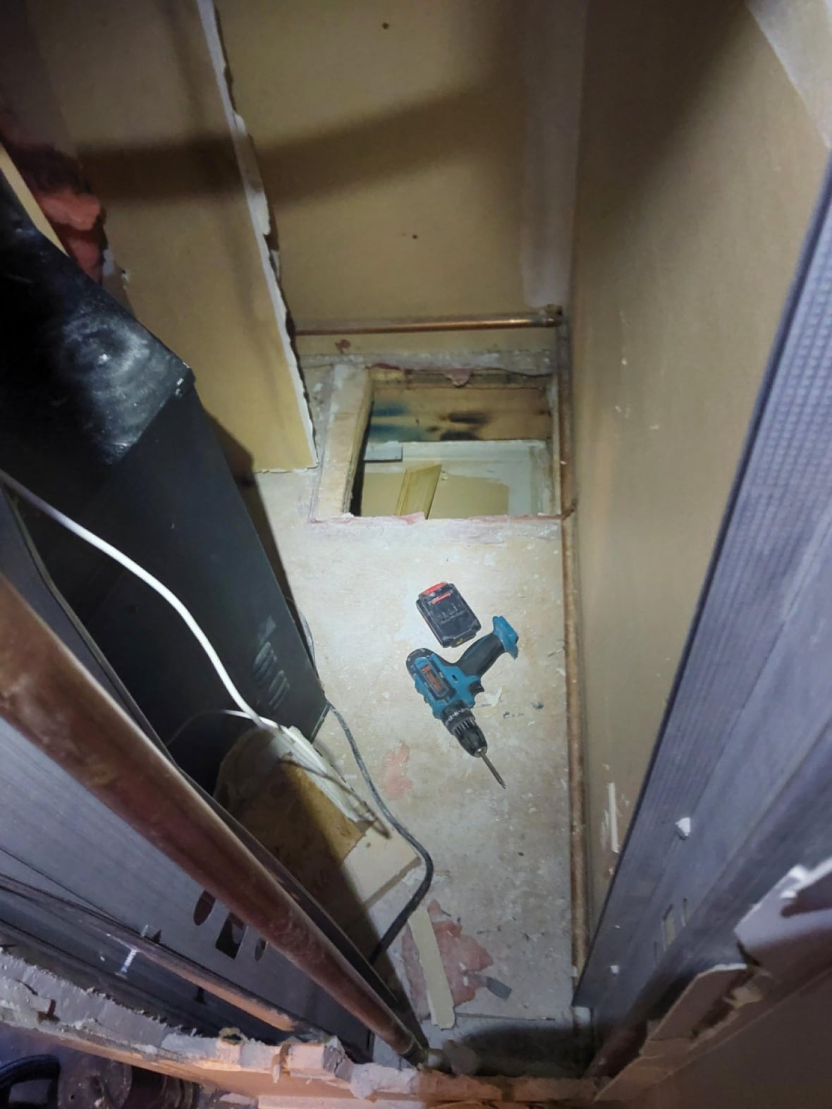 A square shaped hole in the ground with a drill lying on the floor beside it 