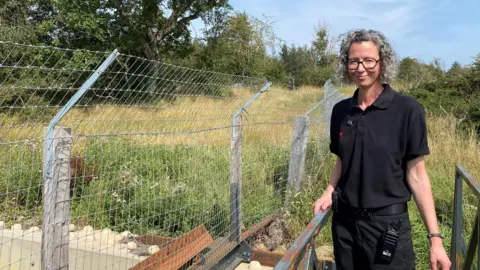 Helen McDonald is standing on a bridge next to a wire fence. She is slim, in her late 30s, wearing a black top and trousers with greyish blonde curly hair and glasses. Trees and long, wild grass sit behind her.