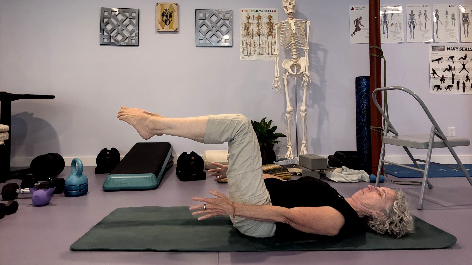 woman in light trousers and dark top lying on the floor legs up at 90 degrees on a green mat in a studio setting.