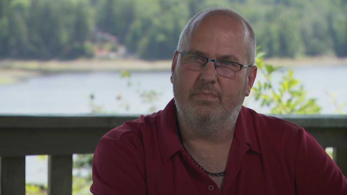 A man wearing glasses, a red polo shirt and black necklace sits in front of a lake. 