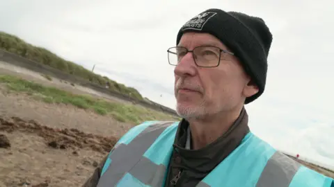 Ras Thomasen is wearing a blue high-vis jacket and a black hat. He is standing on the sand at Morecambe Bay.