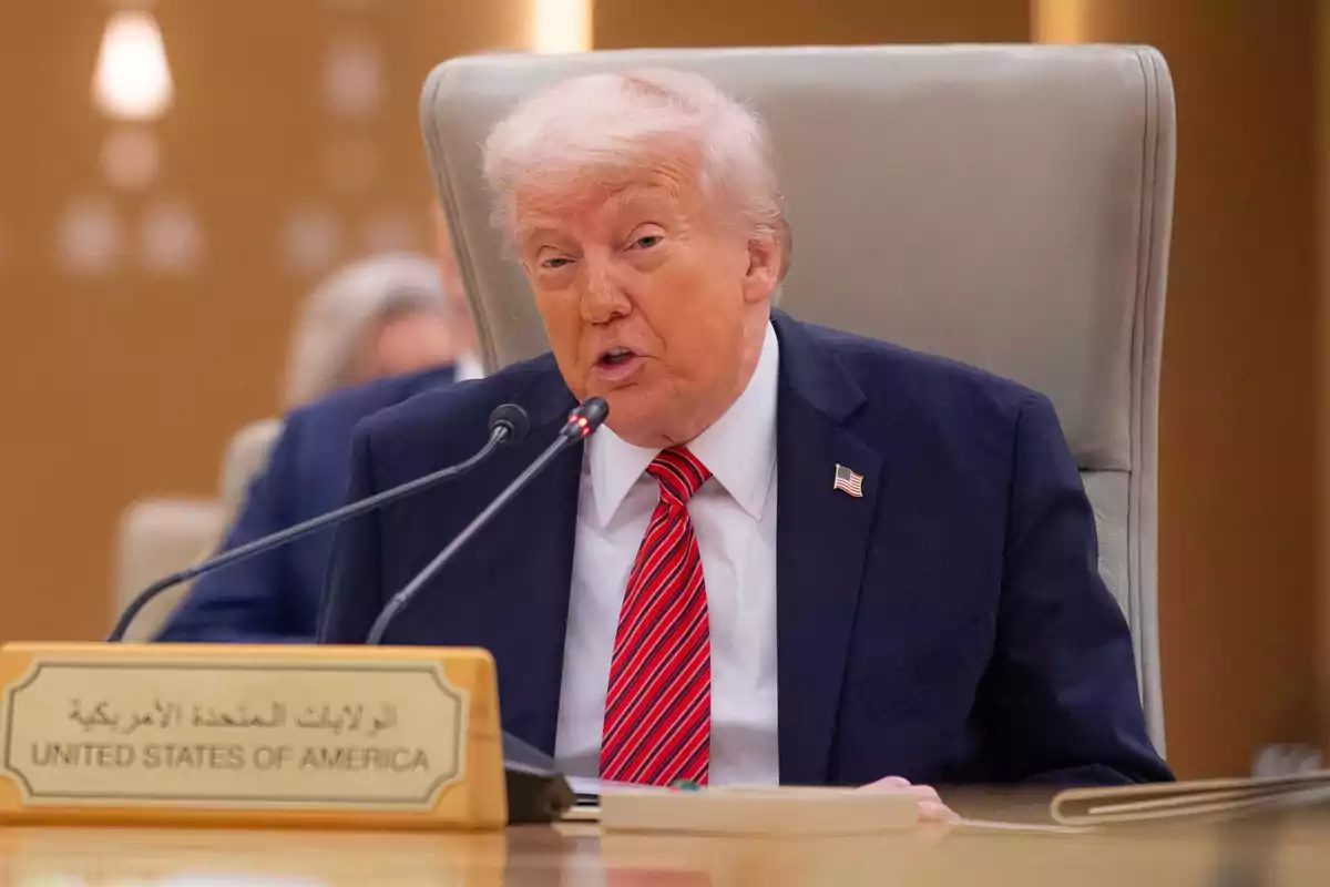 Donald Trump in a dark suit and red tie speaks into a microphone during a conference, with a sign that says Donald Trump in a dark suit and red tie speaks into a microphone during a conference, with a sign that says