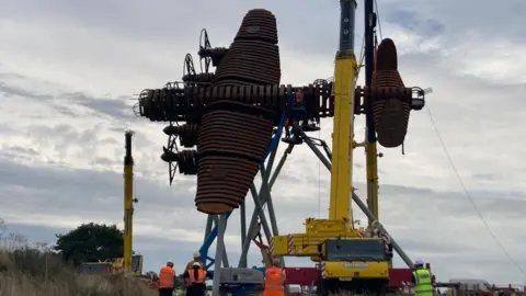 BBC/Jake Zuckerman A large rusted steel structure, in the shape of a World War Two bomber aircraft, is lifted on to a steel support by a large yellow crane as workers in orange and yellow hi-vis jackets watch from the ground. The afternoon sky is darkening.