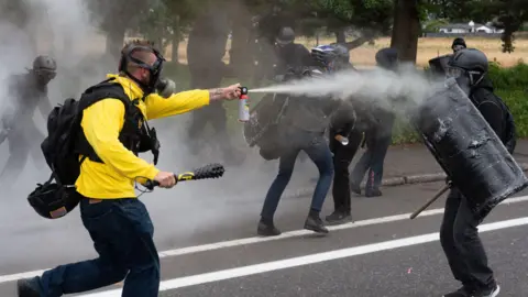 Getty Images Members of the Proud Boys and Antifa clash in Portland, Oregon. A man wearing a gas mask and a yellow jacket sprays a cannister towards a black clad man holding a shield. 