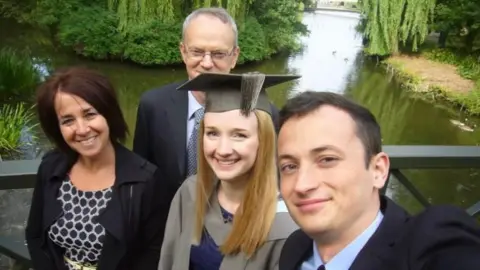 Andrea Brady Jessica Brady wears a university gown and mortar board, surrounded by her family - mum on the left and father standing behind - against a background of pond and trees. All are dressed smartly and are smiling in a selfie.